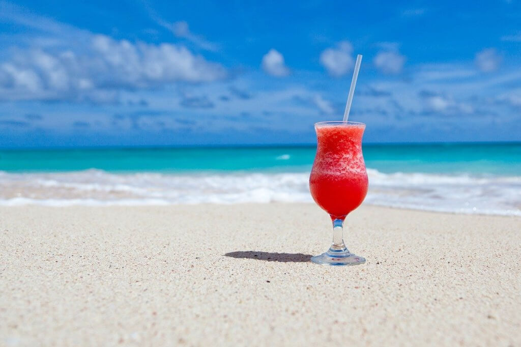 A cocktail on the white sands of a beach; the clear blue ocean on the background merges with a cerulean sky dotted by puffy clouds.