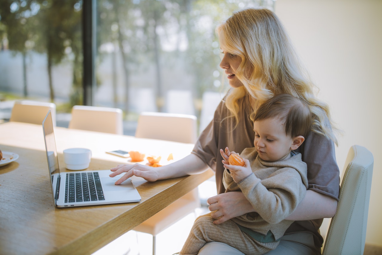 A blonde woman works on her laptop while she holds a baby.