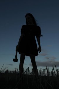 A woman stands against he backdrop of the last light of dusk while she holds an hatchet in her hand.