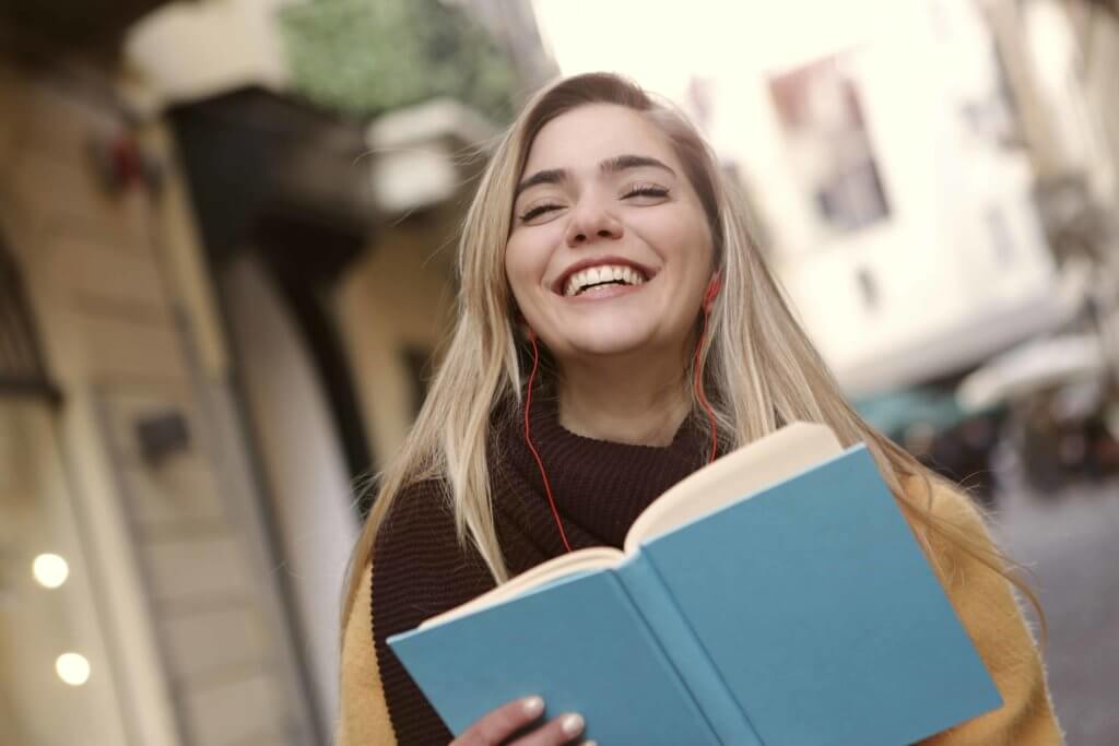 A young woman holds a blue book and laughs with her eyes closed. 