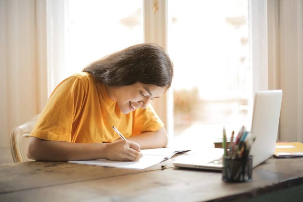 A young woman sits at a desk and writes on a notebook