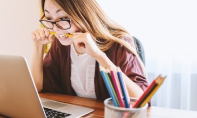 long-haired woman with glasses bites a pencil as she looks at a laptop screen
