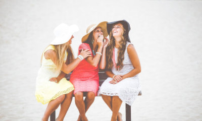 three women wearing hats and summer dresses sitting on a bench talk and laugh amongst themselves